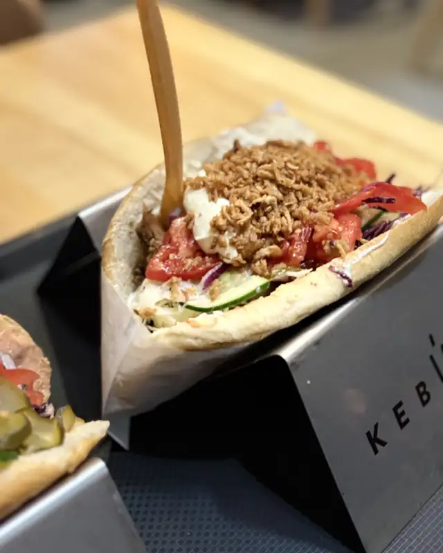 Kebab w bułce z pomidorem i prażoną cebulką na czarnym stojaku z napisem KebUp